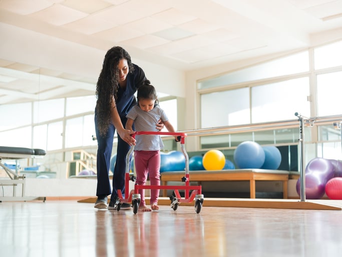 A physical therapist helping a little girl with autism to walk with a wheeled walking frame