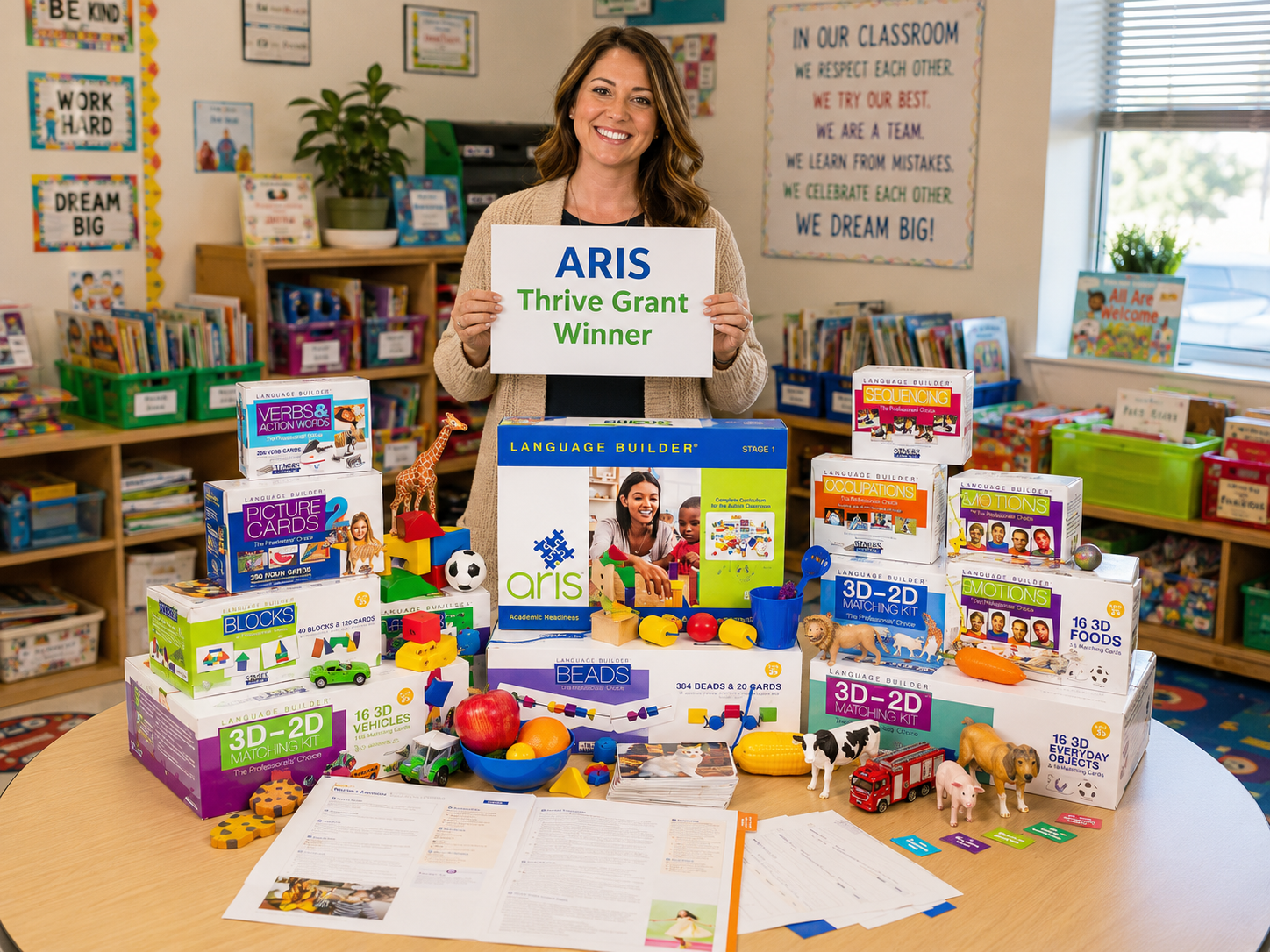A smiling ARIS Thrive Grant–winning teacher in her bright classroom holding an 'ARIS Thrive Grant Winner' sign, with the full ARIS curriculum kit spread on the table in front of her