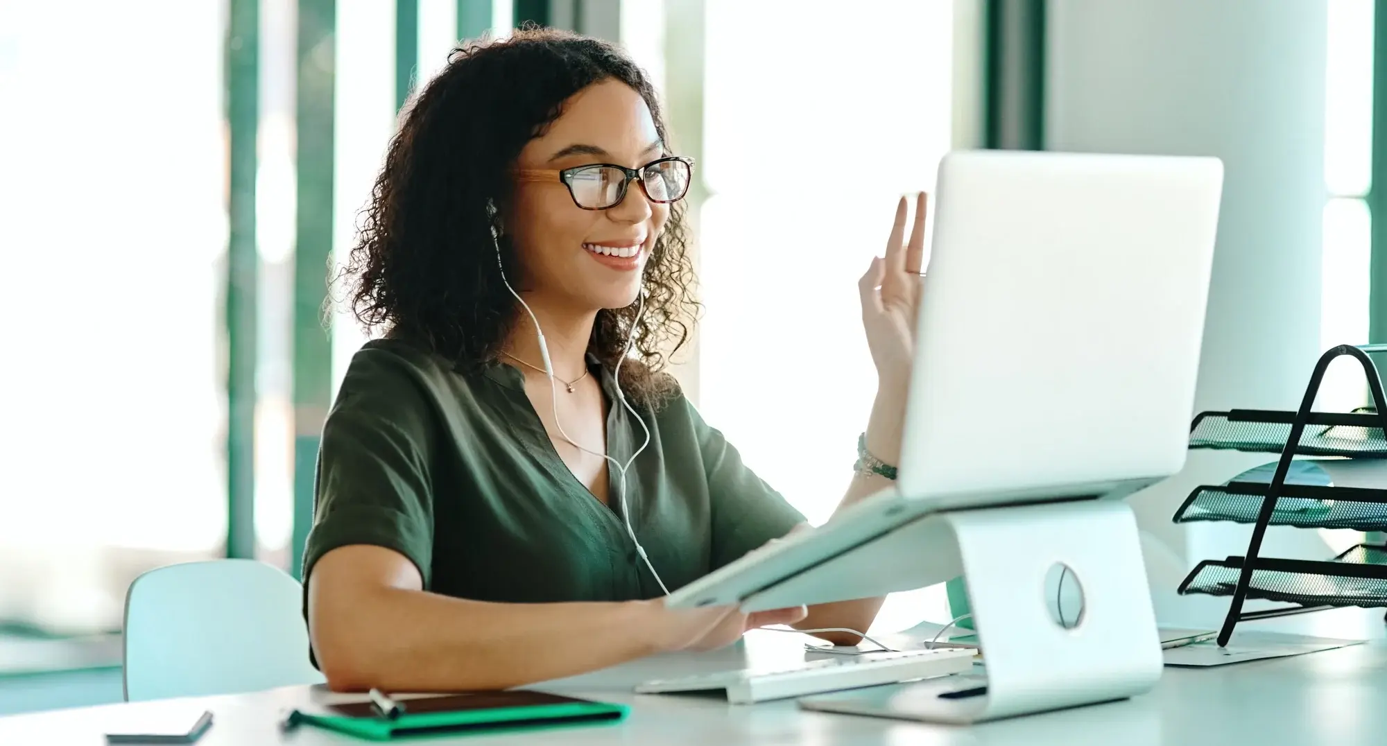 Woman with glasses and earbuds waving at laptop screen during video call at desk