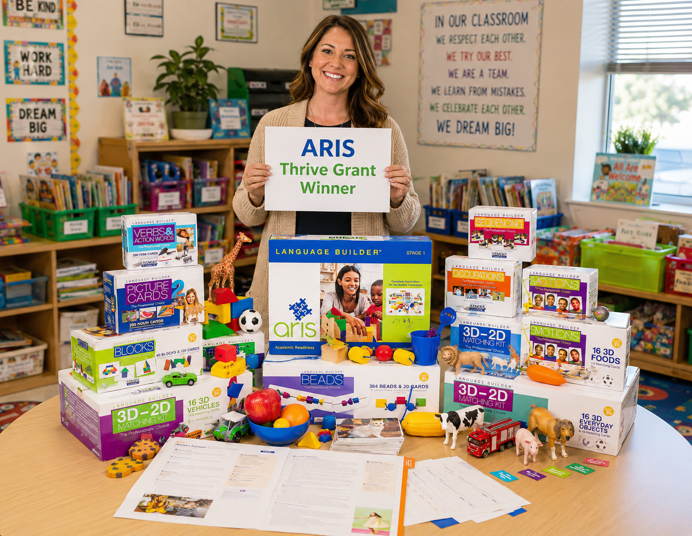 A smiling ARIS Thrive Grant–winning teacher in her bright classroom holding an 'ARIS Thrive Grant Winner' sign, with the full ARIS curriculum kit spread on the table in front of her